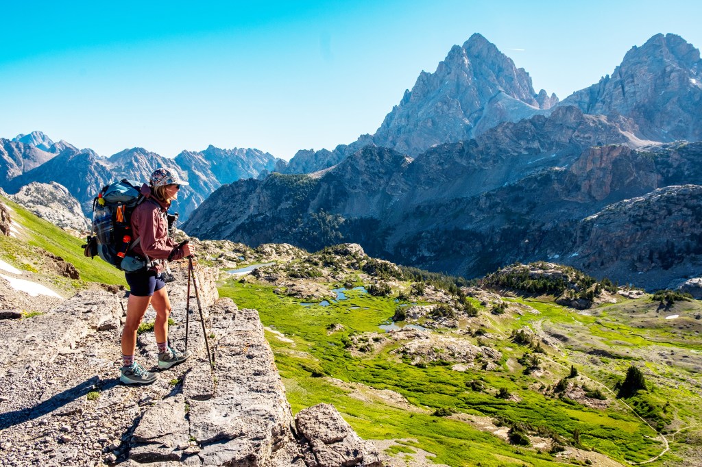 A hiker stands on a rocky ledge, overlooking a lush green valley with mountains in the background on a sunny day.