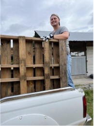 A person smiling while balancing on the edge of a wooden pallet in the bed of a pickup truck, set against a cloudy sky and a farm backdrop.