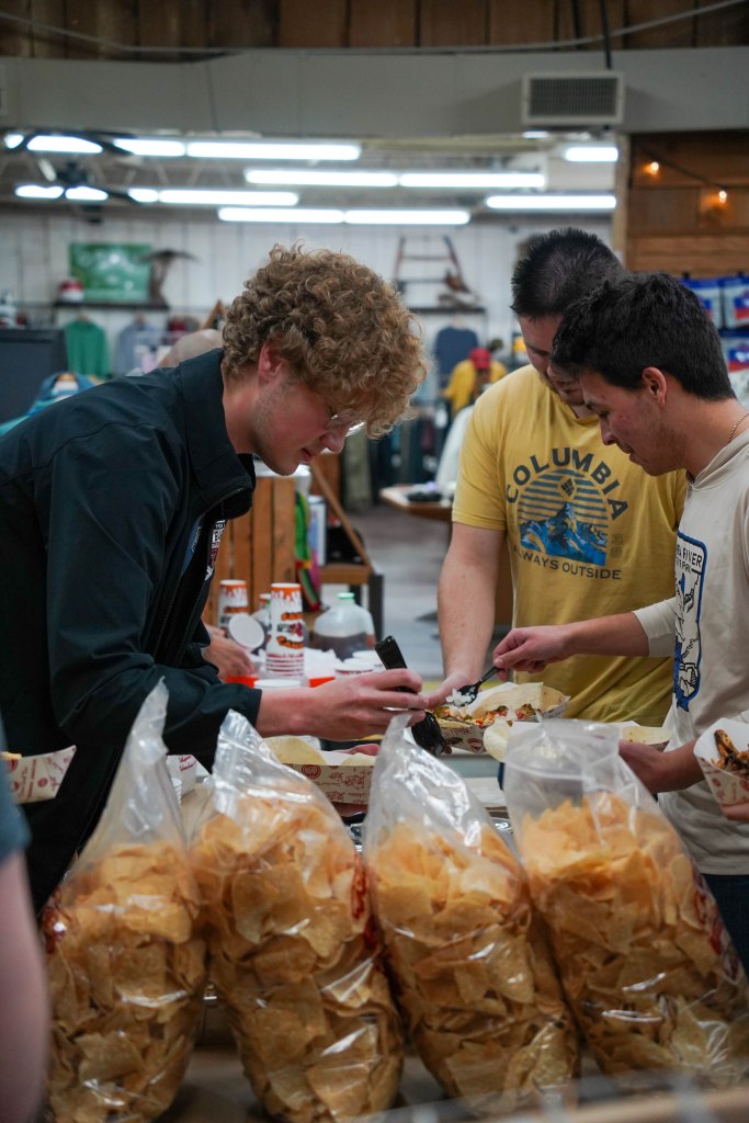 A group of young men interacting at a food table, one is serving food while others are preparing to receive it. Bags of chips are visible in the foreground.