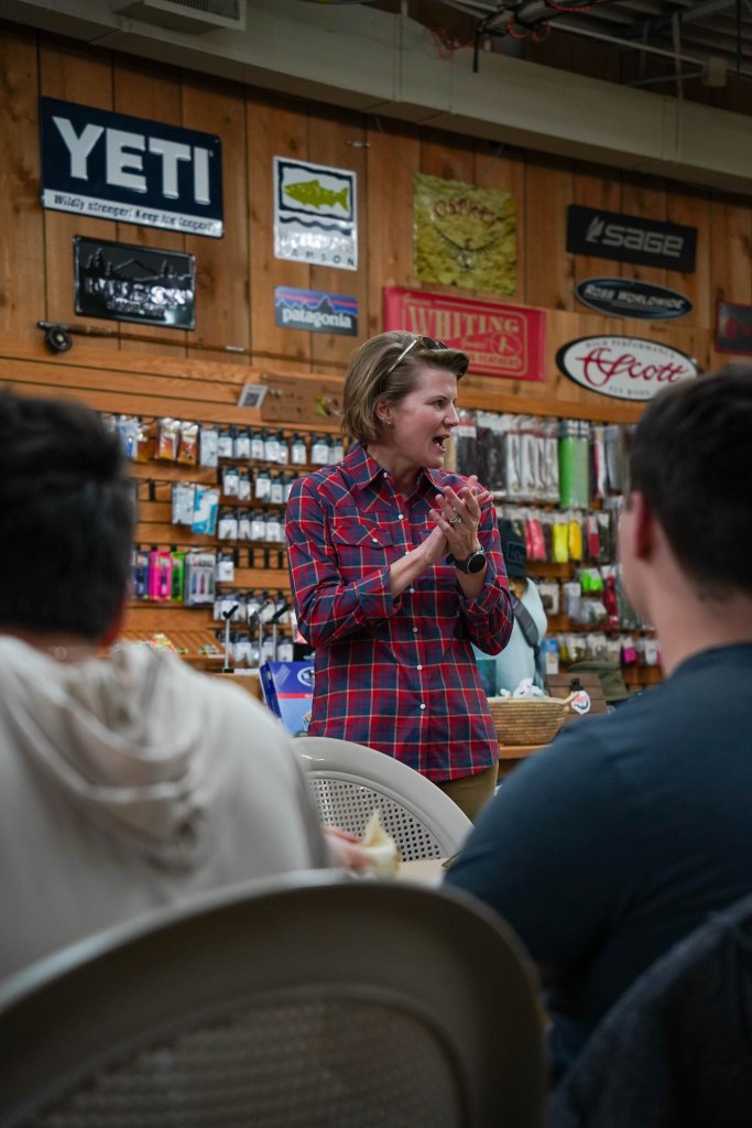 A person in a plaid shirt is speaking to a group in a retail store filled with outdoor gear and apparel displays.