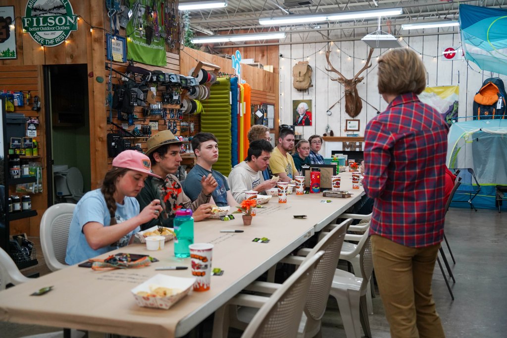A group of students seated at a long table in an outdoor gear store, eating and listening to a speaker. The setting includes various camping and hiking supplies displayed on the walls.