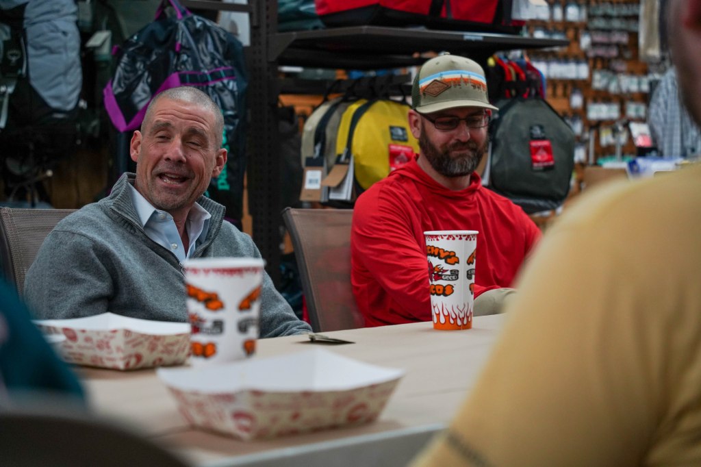 Two men seated at a table inside a gear shop, one wearing a grey sweater and the other in a red hoodie, engaging in discussion.