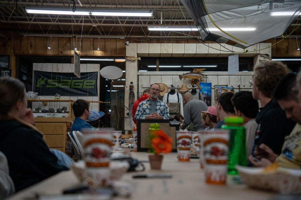 A speaker addressing a group of students inside a rustic outdoor gear shop, with various equipment and posters displayed in the background.