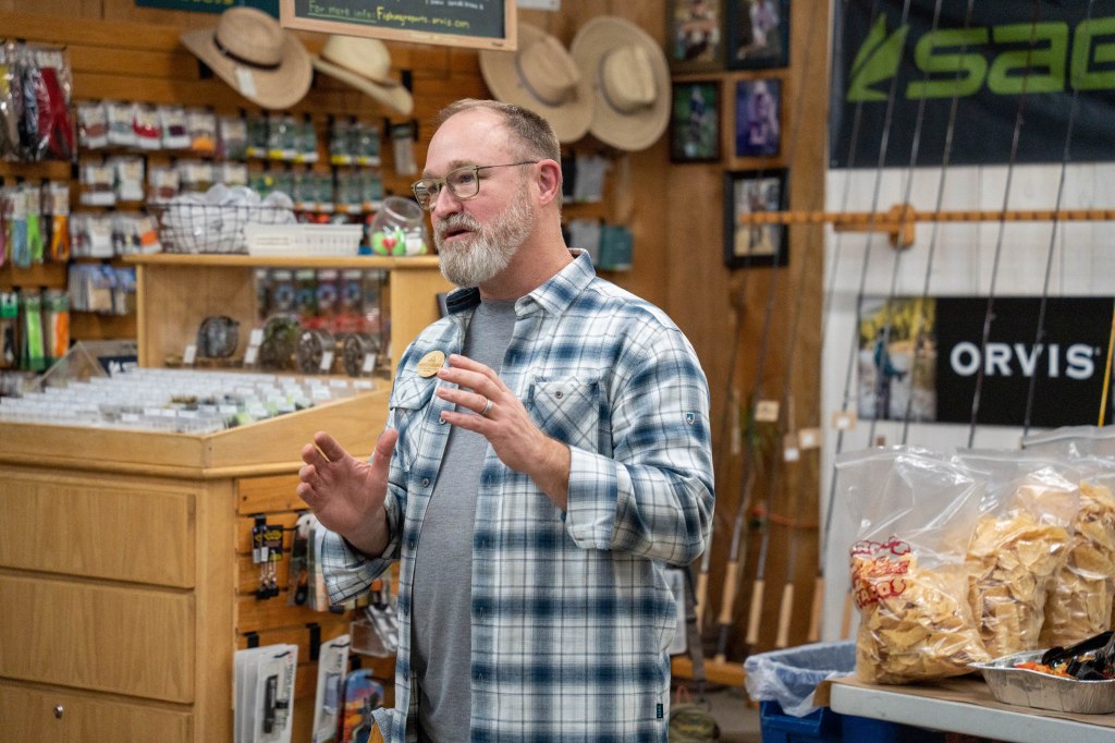 A speaker with glasses and a beard gestures while giving a presentation in a store filled with outdoor gear and supplies.