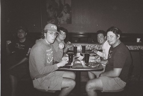 A group of five young men sitting at a table in a dimly lit restaurant, enjoying food and drinks together.
