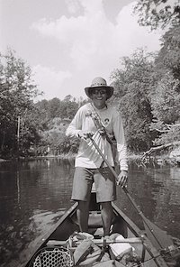 A person paddling a canoe on a river surrounded by lush greenery, wearing a wide-brimmed hat and sunglasses.