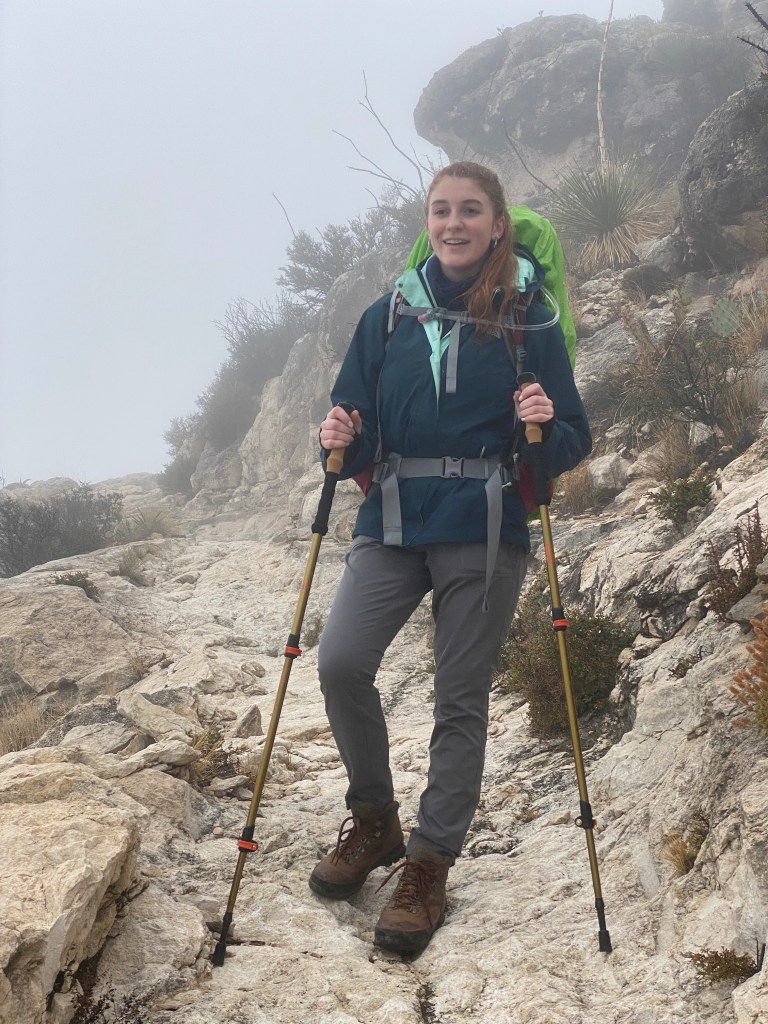 A student hiking on a rocky trail in foggy conditions, wearing a blue jacket and carrying trekking poles, in Guadalupe Mountain National Park.