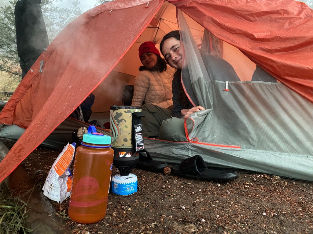 Two students sitting inside a tent during a camping trip. A camping stove and a water bottle are visible in the foreground.