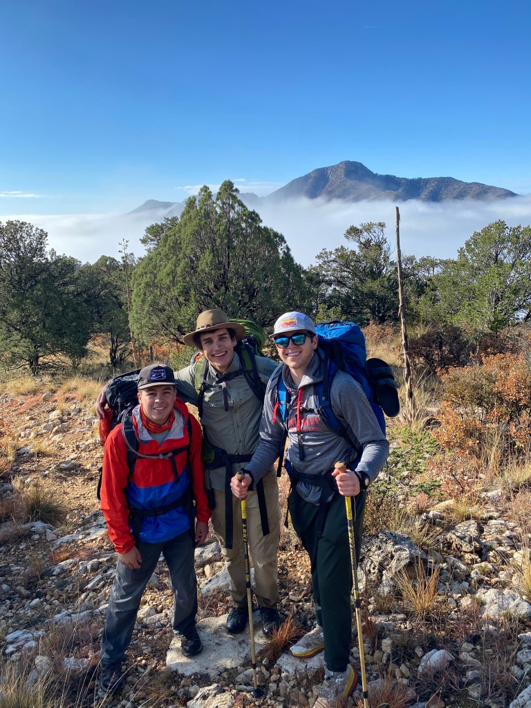 Three male students pose together on a hiking trail in Guadalupe Mountains National Park, surrounded by trees and misty mountains in the background.