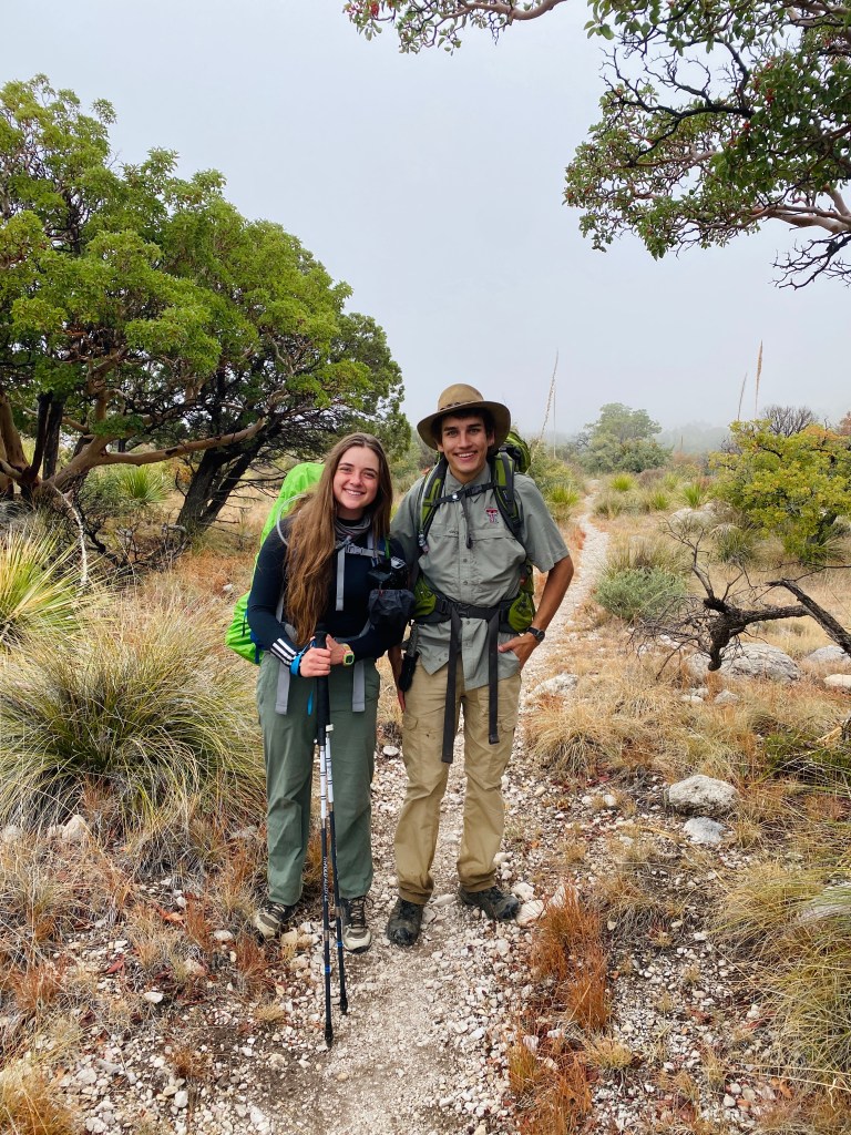 Two students smiling on a hiking trail in Guadalupe Mountain National Park, surrounded by vegetation and fog, with backpacks and trekking poles.