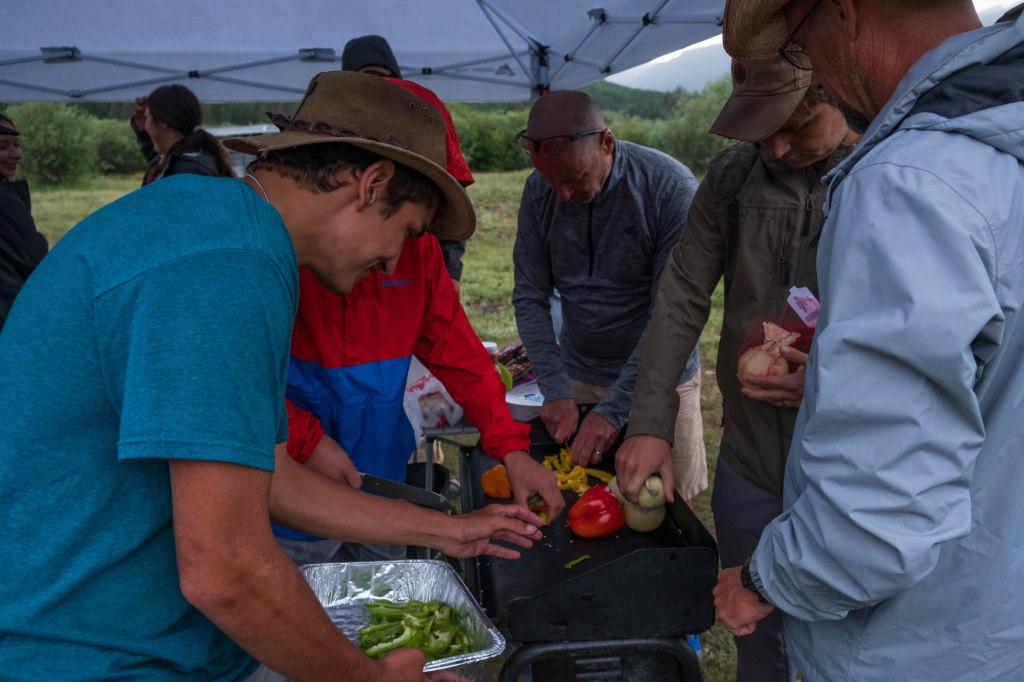 A group of individuals preparing vegetables at a campsite kitchen under a tent, with mountains and greenery in the background.