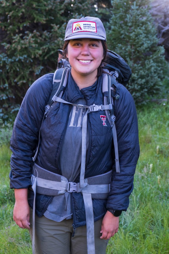 A smiling female student wearing a backpack and a 'Bridge Adventure' cap, standing in a grassy area with trees in the background.