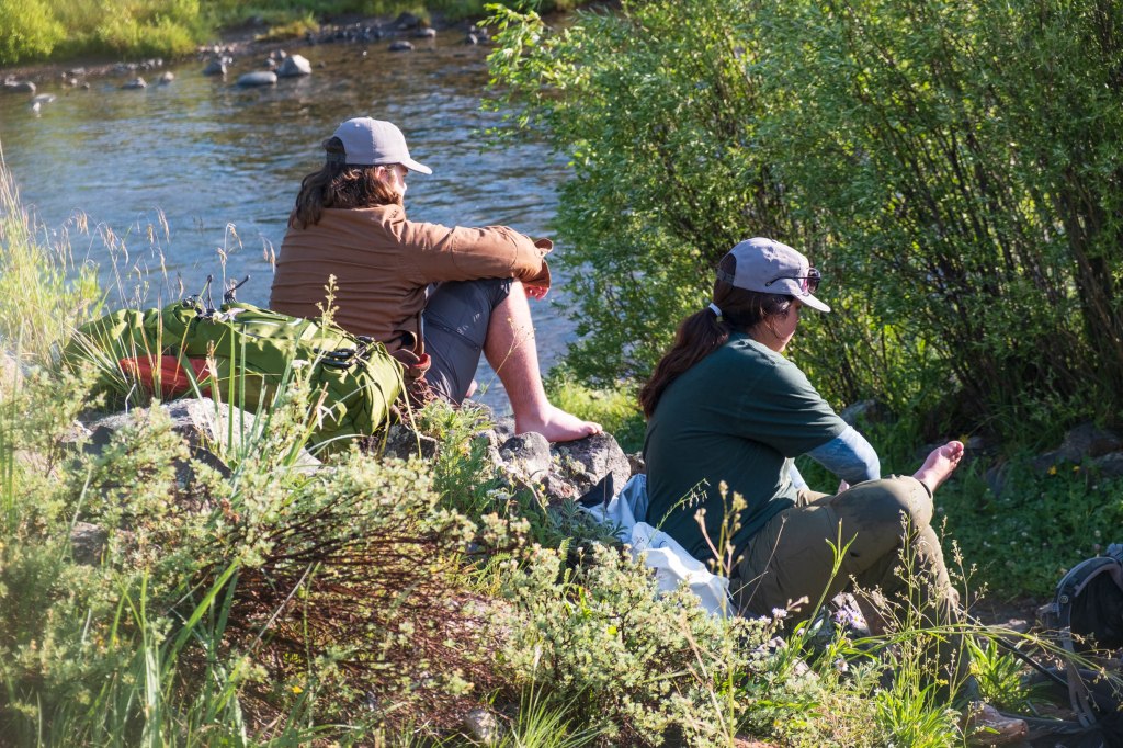 Two individuals sitting by a river, surrounded by greenery and rocks. One is wearing a brown jacket and the other is in a green shirt, both appear to be resting.