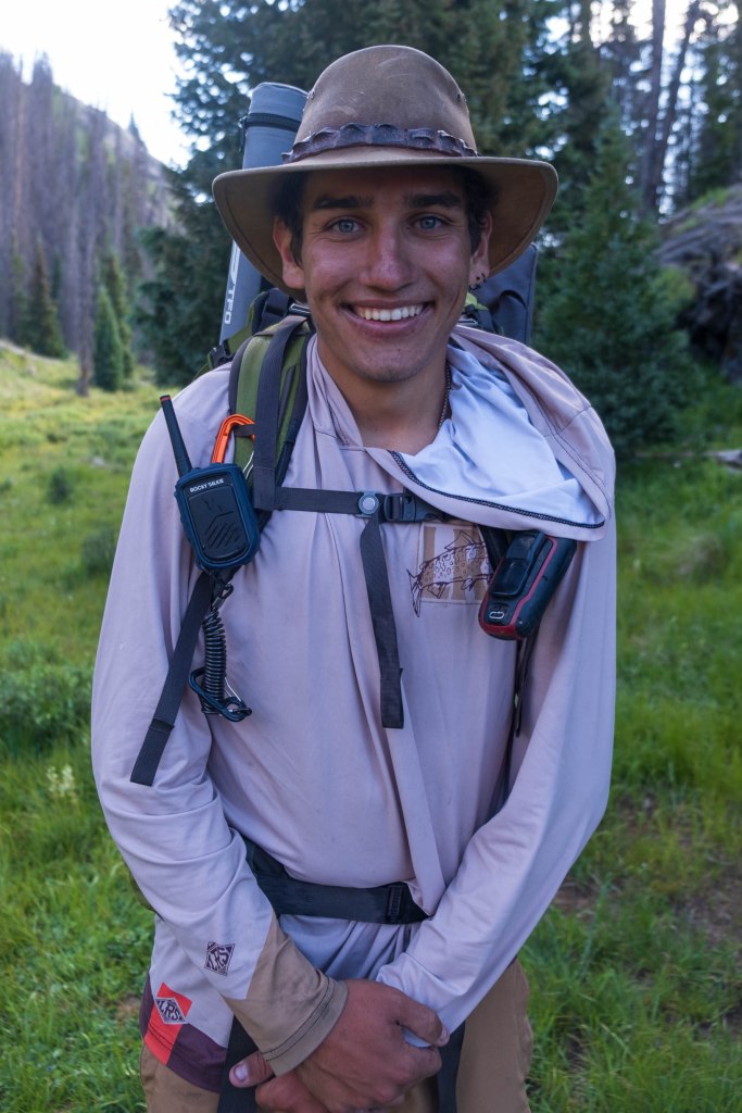 A young man with a wide smile, wearing a hat and outdoor gear, standing in a grassy area surrounded by trees.