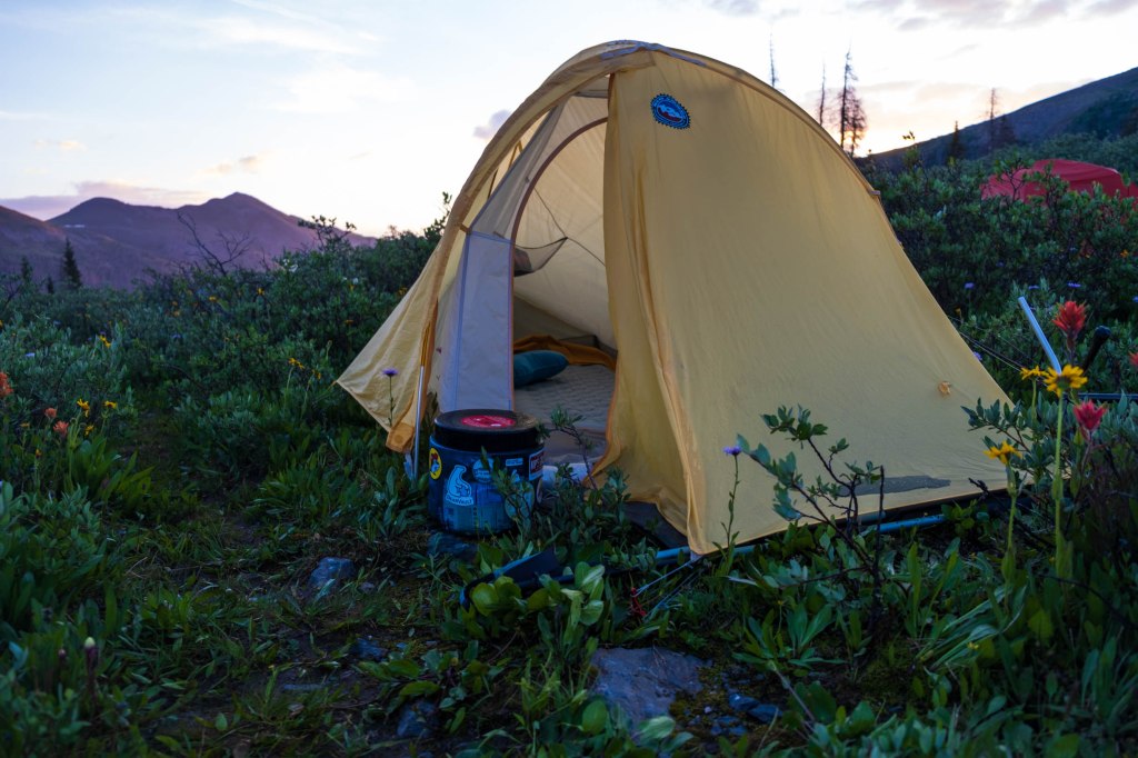 A yellow tent set up in a grassy field with blooming wildflowers, surrounded by mountains during twilight.