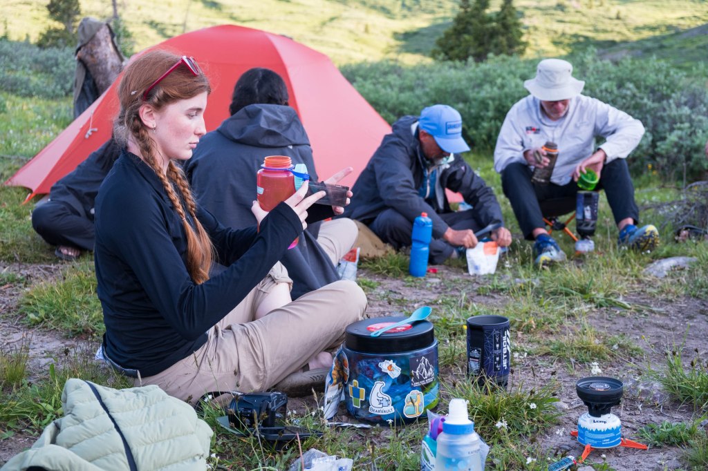 A group of people preparing food at a campsite in the wilderness, with a red tent in the background.