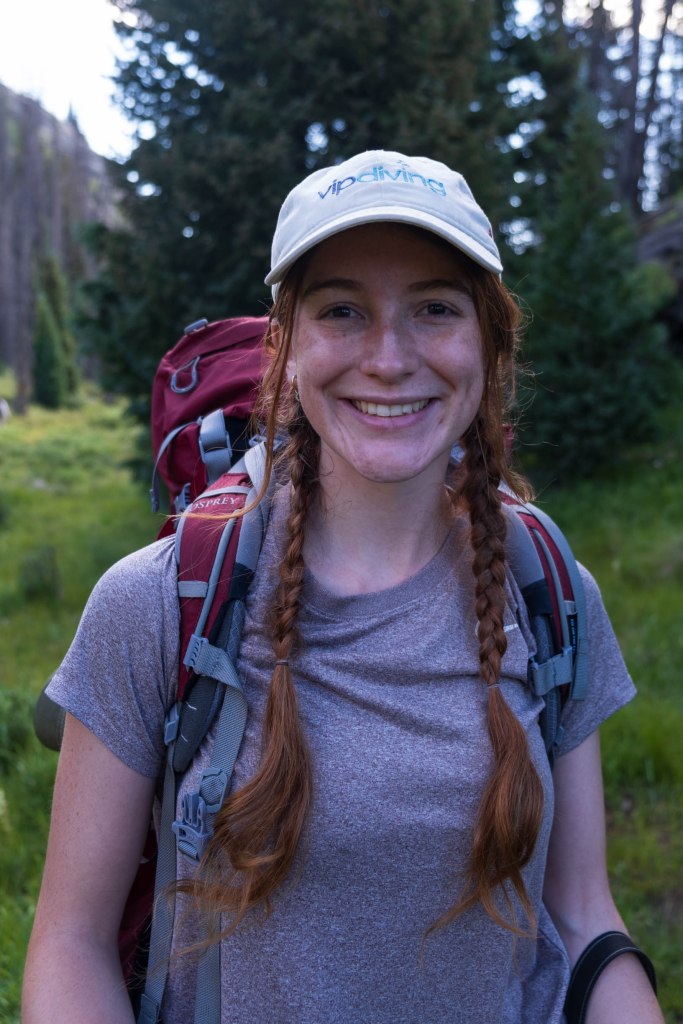 A smiling female hiker with long brown braids wearing a gray t-shirt and a white cap stands in a lush green forest setting, carrying a red backpack.