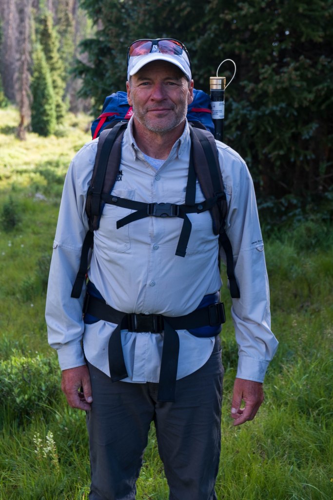 A man wearing a light-colored long-sleeve shirt and a cap stands in a green, outdoor setting with a backpack.