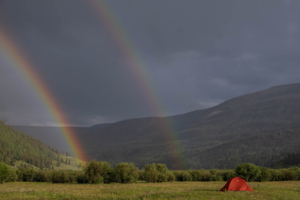 A vibrant scene showing a red tent in a grassy field under a cloudy sky with two rainbows arching over the mountains.