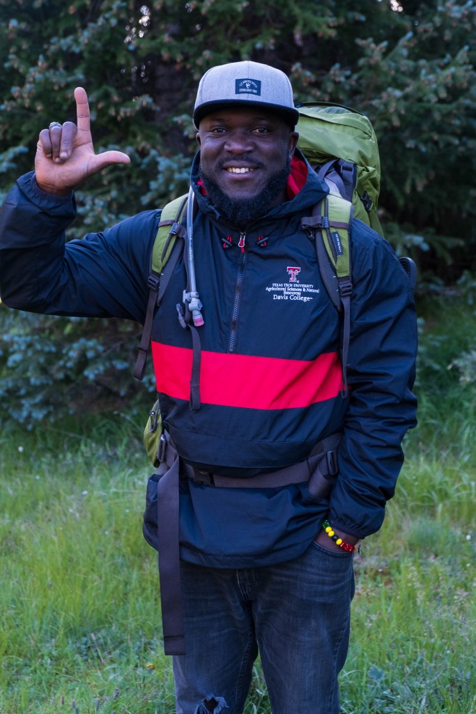 A smiling individual wearing a black and red jacket, cap, and a backpack, standing in a grassy area and making a hand gesture.