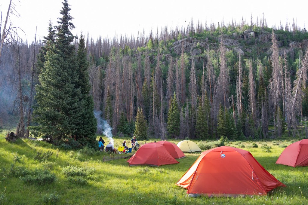 A camping scene in the Weminuche Wilderness featuring red tents set up on a grassy area with a group of people gathered around a fire in the background, surrounded by pine trees and mountain terrain.