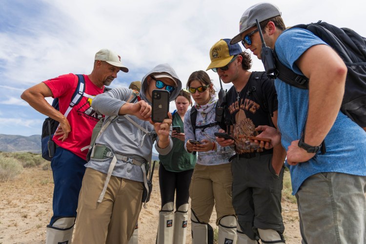 A group of students and faculty members from Texas Tech University's Bridge Adventure Program gather outdoors, using their phones to examine something together, while wearing outdoor gear.