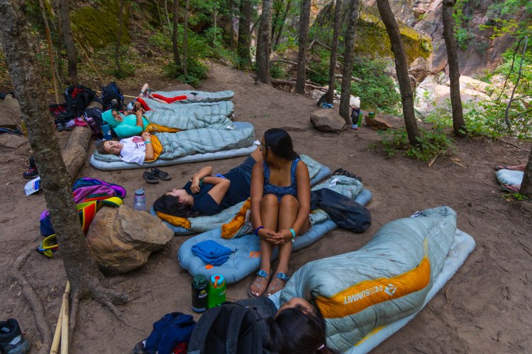 Students relaxing on sleeping pads in a forested campsite during the Bridge Adventure Program in Zion National Park.