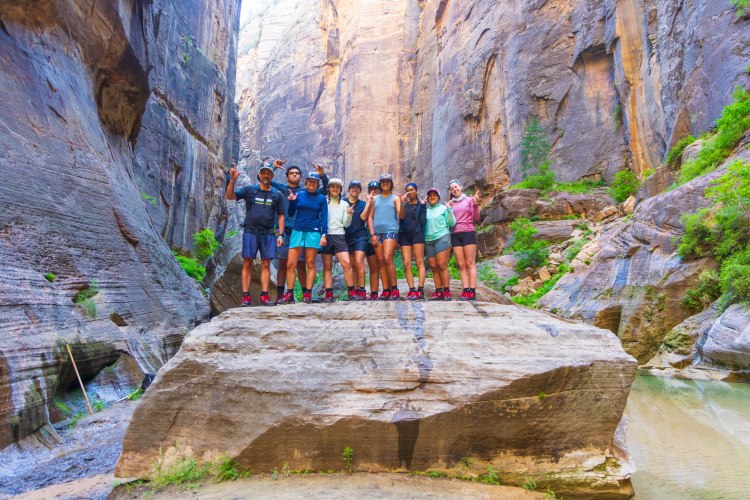 Group of students and faculty posing on a large rock in the Narrows of Zion National Park, surrounded by towering canyon walls.